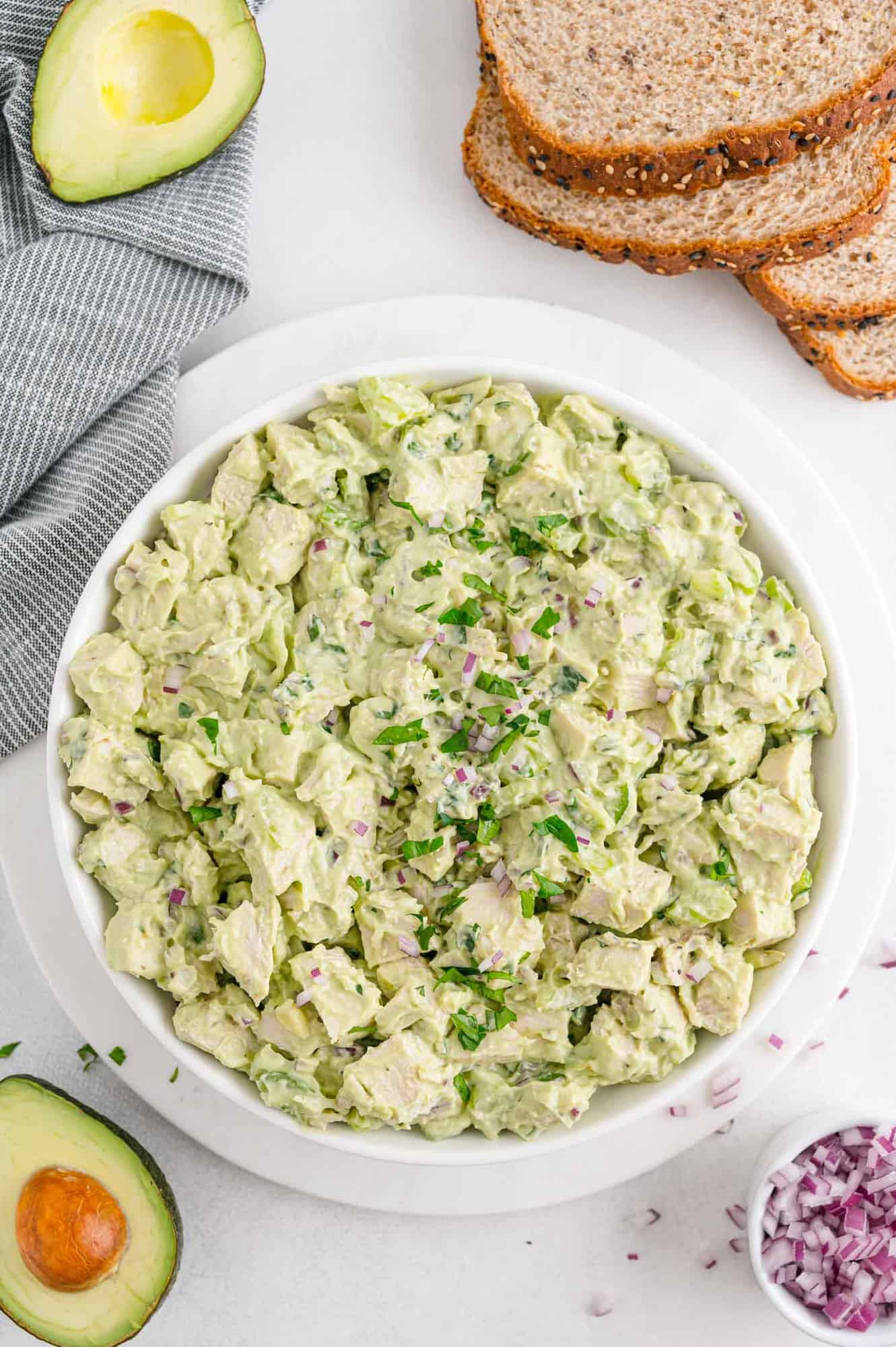 Avocado chicken salad in a bowl, surrounded by bread, avocados, onion.