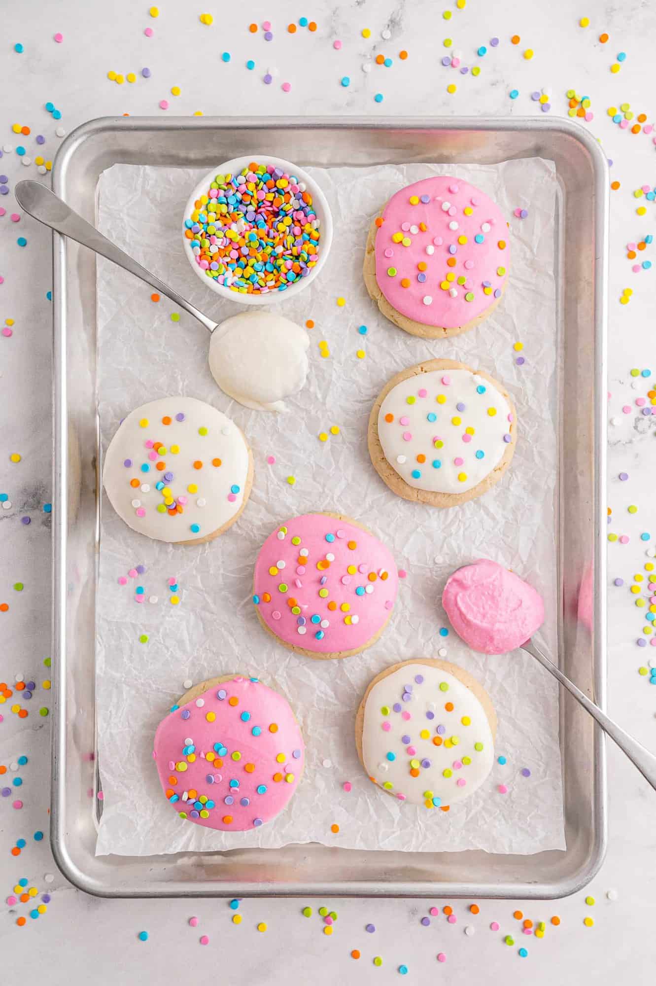 Tray of cookies with spoons full of frosting and a bowl of sprinkles.