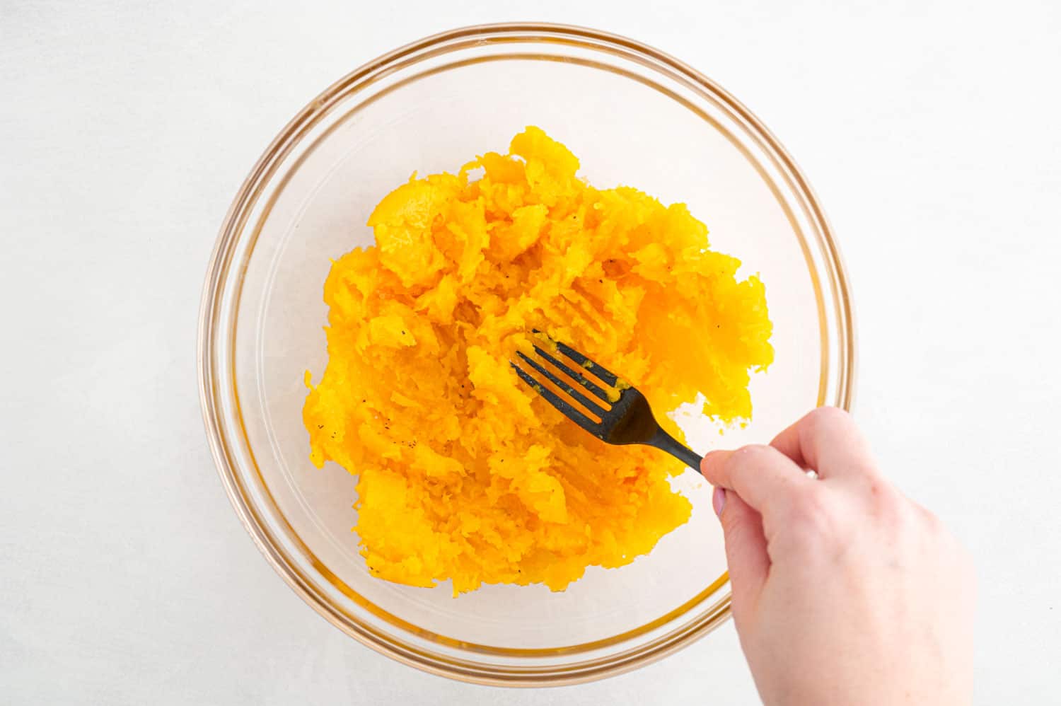 Squash flesh being mashed with a fork in clear bowl.