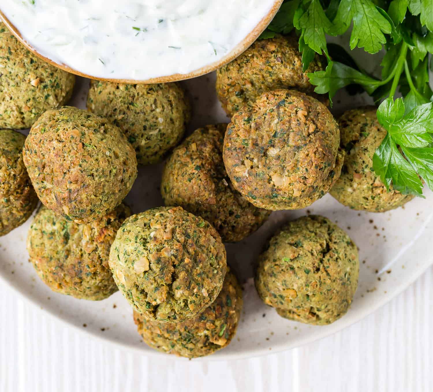 Overhead view of crispy chickpea balls, aka falafel. A bowl of tzatziki sauce and fresh parsley are also partially visible.