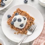 Square of baked oatmeal on a white plate, topped with yogurt and fresh blueberries.