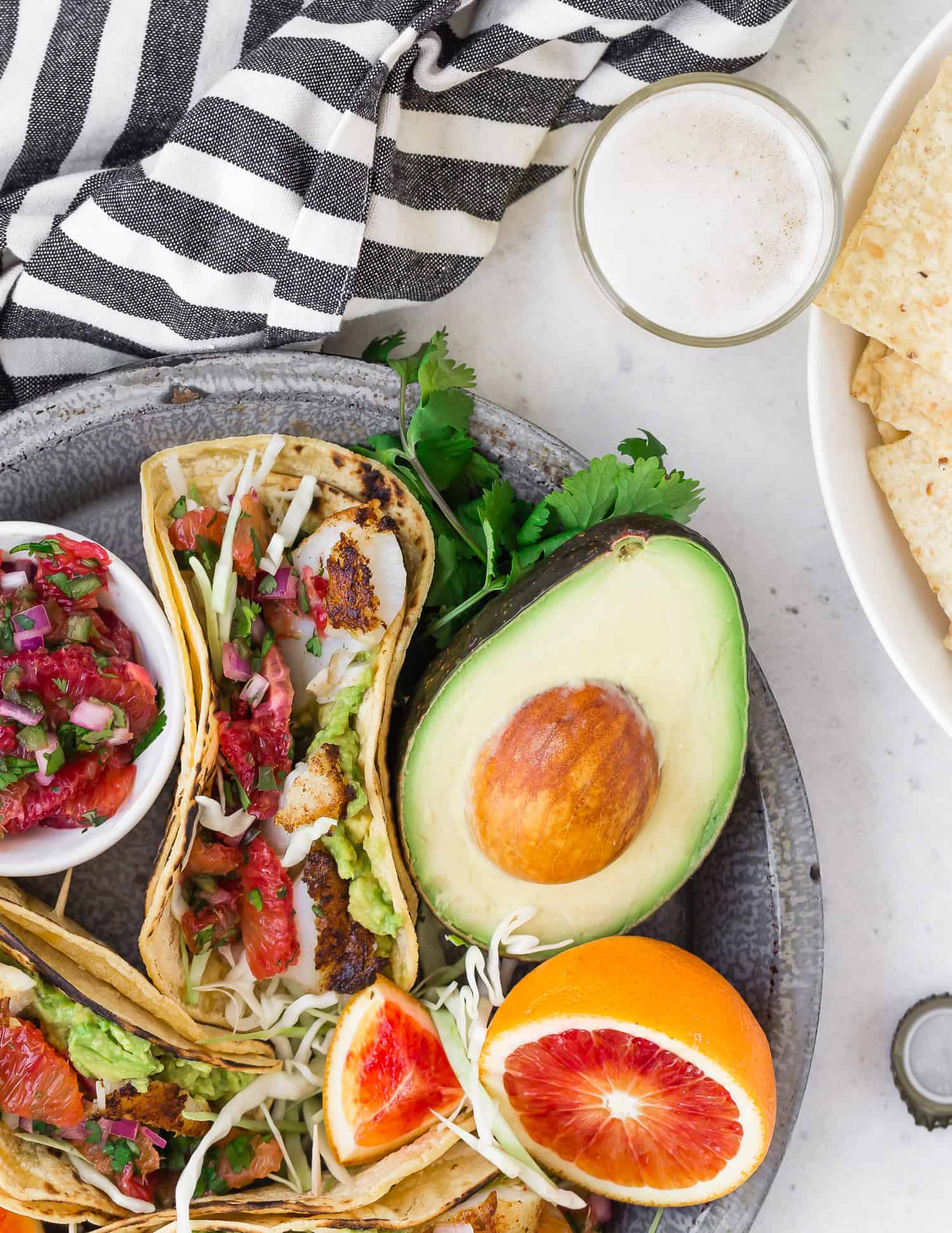 Tacos on a platter with avocado, blood orange, a beer, and chips nearby.