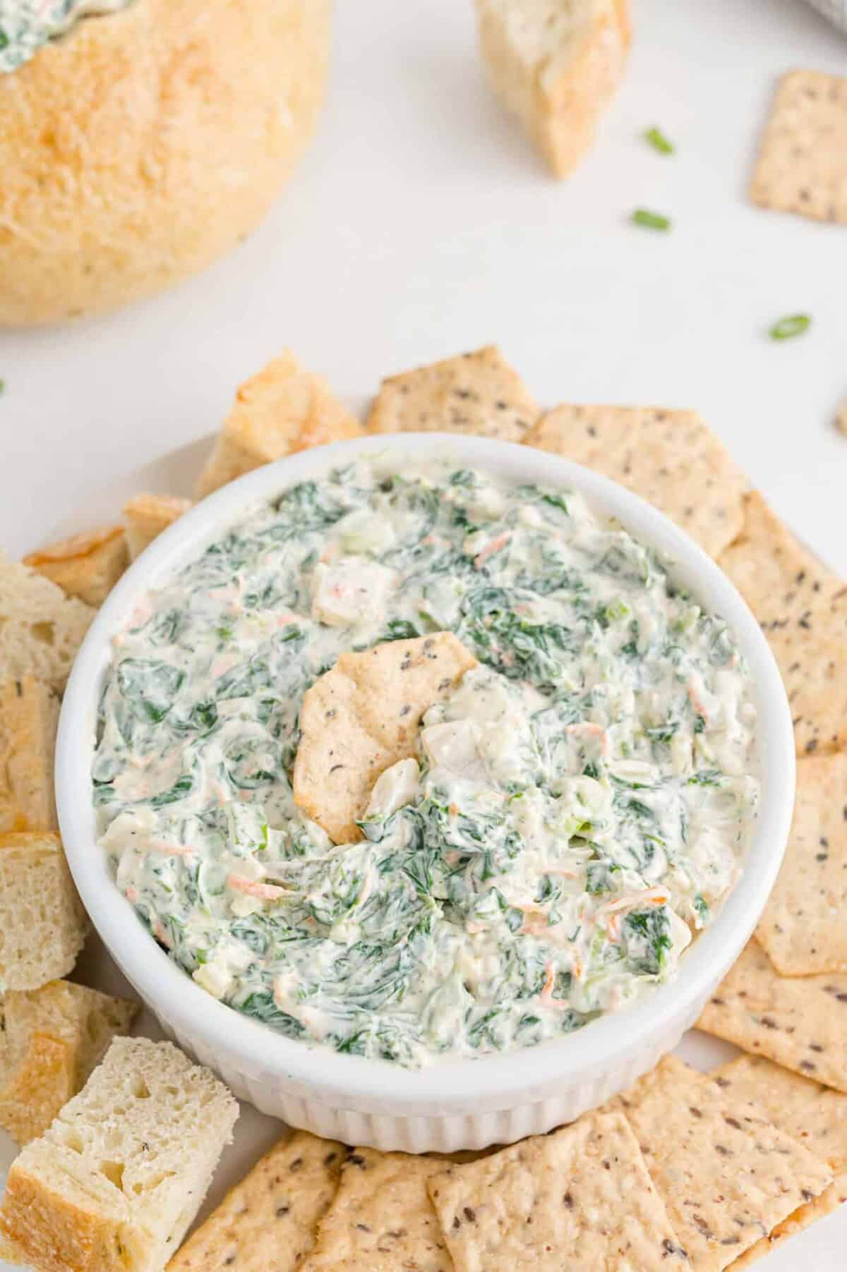 A white bowl filled with creamy spinach dip, surrounded by crackers and bread pieces, sits on a light-colored surface.