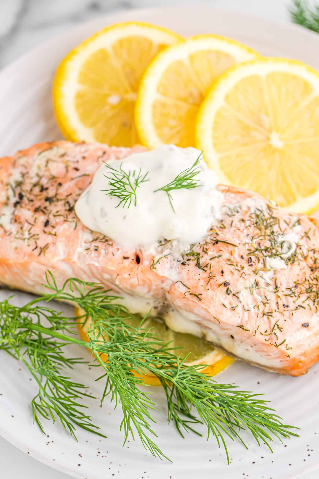 Lemon dill salmon topped with white sauce and fresh dill, next to lemon slices on a plate.