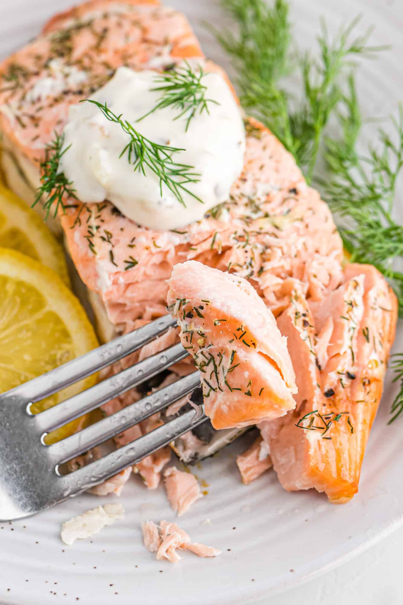 A fork lifting a piece of lemon salmon from a fillet topped with sour cream and dill sprigs.