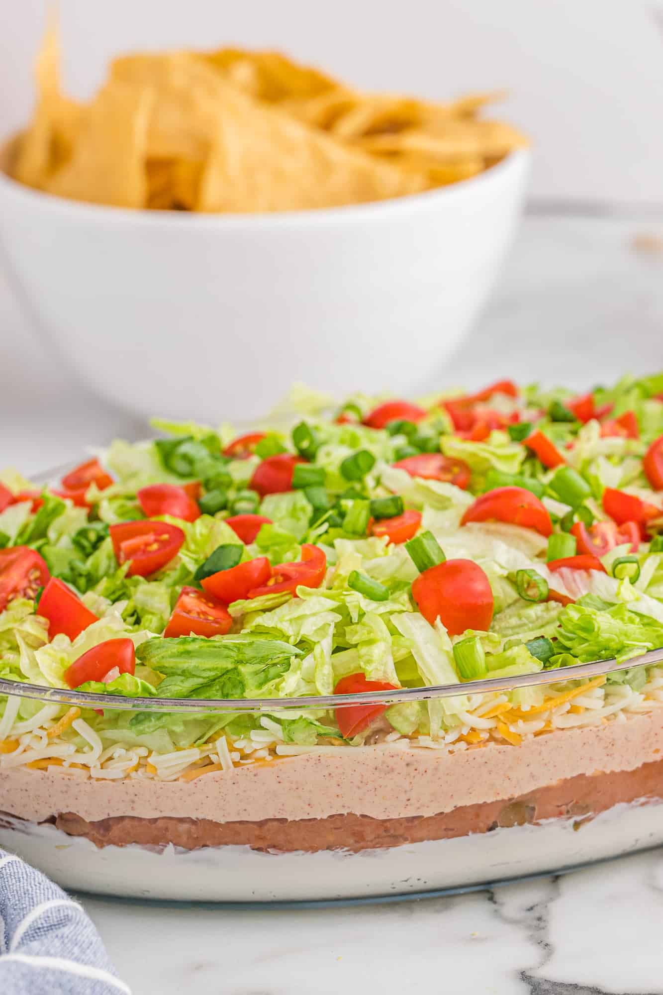 7 Layer dip in a glass pan, bowl of chips in background.