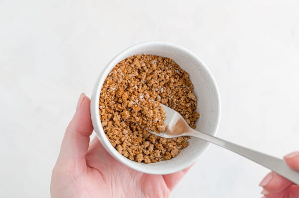 A hand holding a small white bowl with salmon seasoning, while the other hand mixes the spices together with a fork.