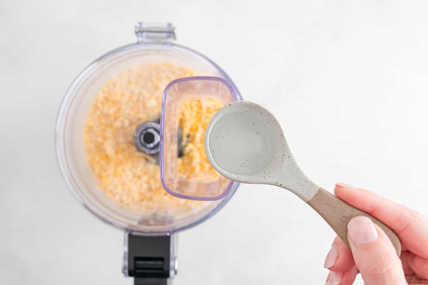A hand holds a ceramic spoon with liquid above a food processor containing crushed ingredients.