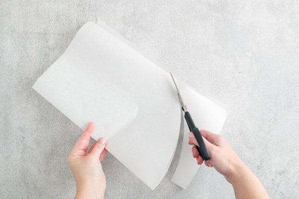 One hand holding folded parchment paper while the other hand uses scissors to cut out a half-oval.