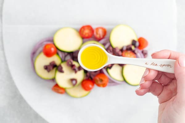 A spoonful of olive oil held over vegetables arranged on a parchment paper oval.