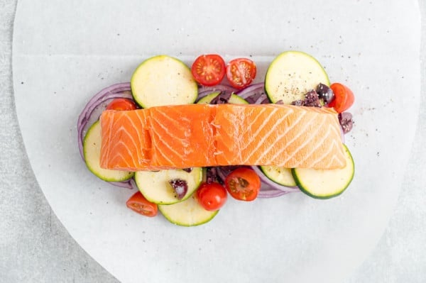 A salmon fillet placed over a bed of vegetables on a parchment paper oval.