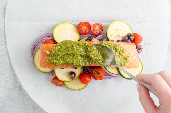 A spoon spreads pesto over a salmon fillet on a bed of vegetables on a parchment paper oval.