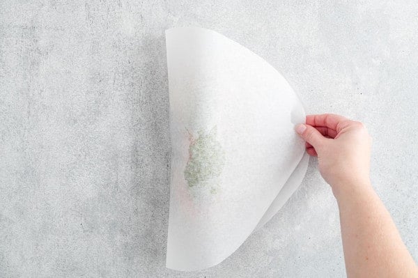 A hand folds a parchment paper oval in half over pesto salmon ingredients.