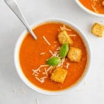 Tomato soup in white bowl garnished with croutons, cheese, basil.