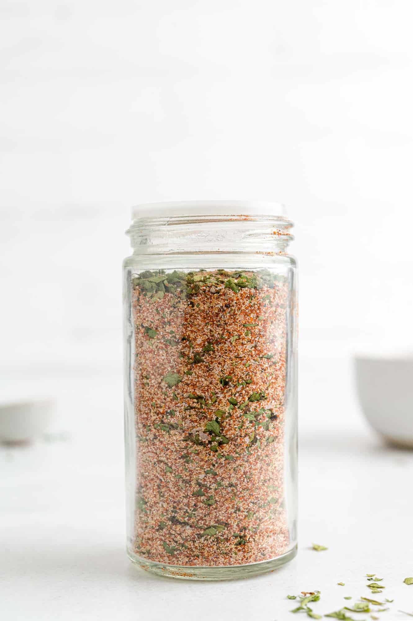 A clear glass jar filled with a coarse spice blend, featuring red and green specks, sits on a white surface with blurred bowls in the background.