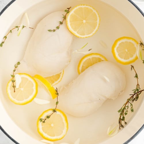 Poached chicken breasts in a pan with water, lemon slices, thyme.