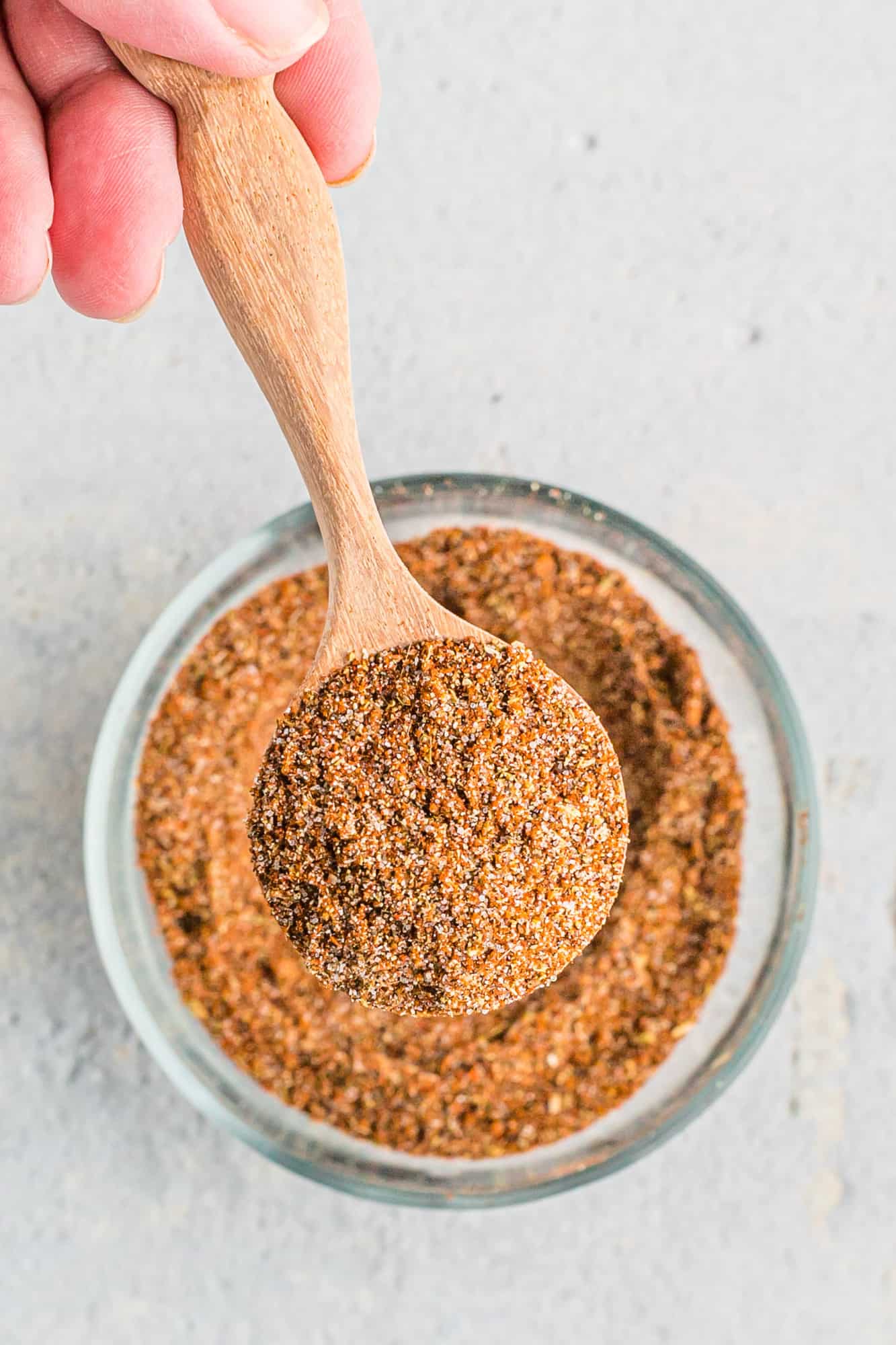 A hand holds a wooden spoon filled with a fajita spice blend above a glass bowl containing more of the same spice mixture.