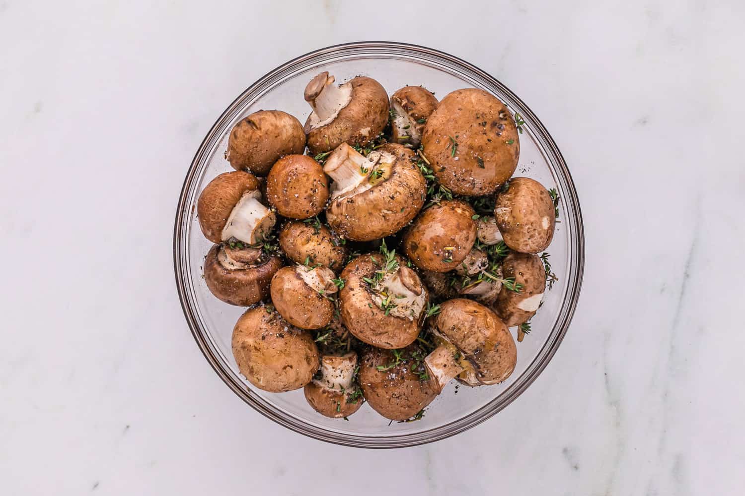 Uncooked mushrooms in glass bowl with olive oil and seasonings.