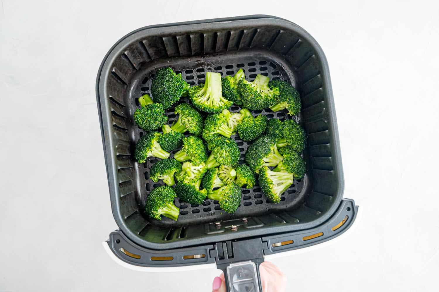 Hand shaking air fryer basket full of broccoli.