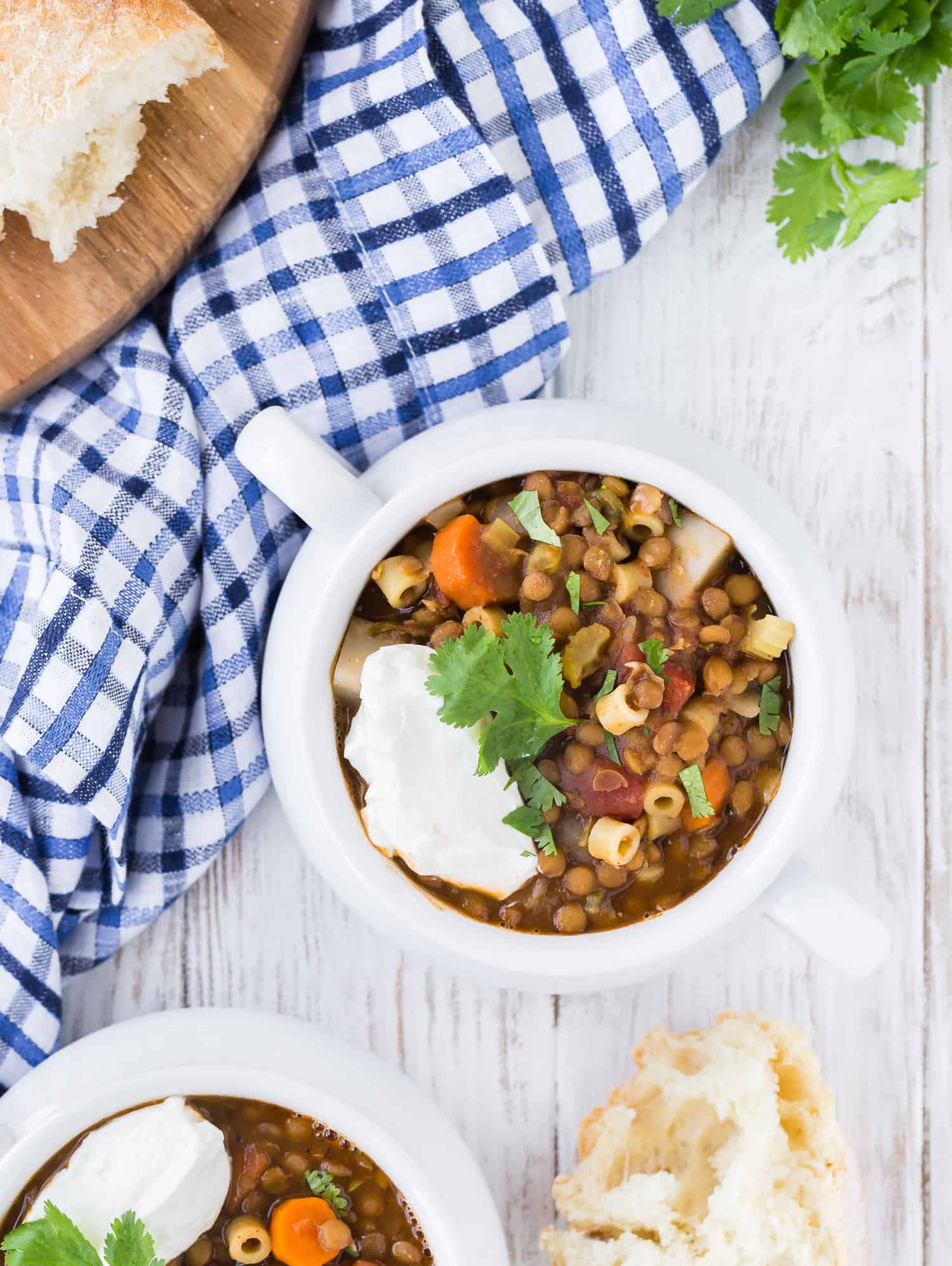 Overhead photo of two bowls of lentil soup, one only partially showing in image. Cilantro is also pictured as well as fresh bread that has been torn from the loaf. 