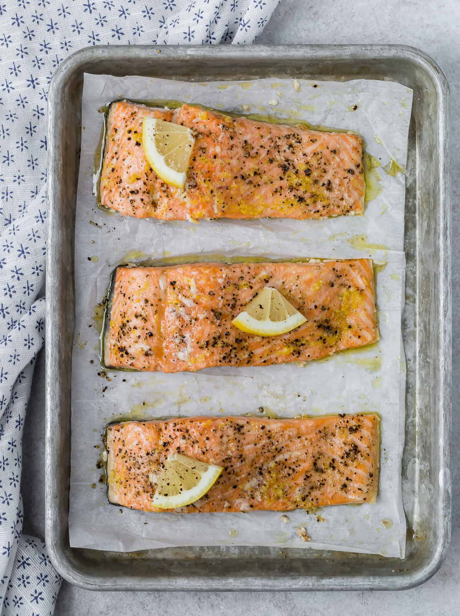 Overhead view of three salmon filets on a baking sheet topped with lemon slices and black pepper.