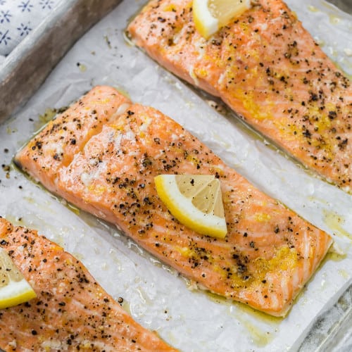 Three slow roasted salmon filets topped with lemon slices and black pepper on a parchment lined baking sheet.