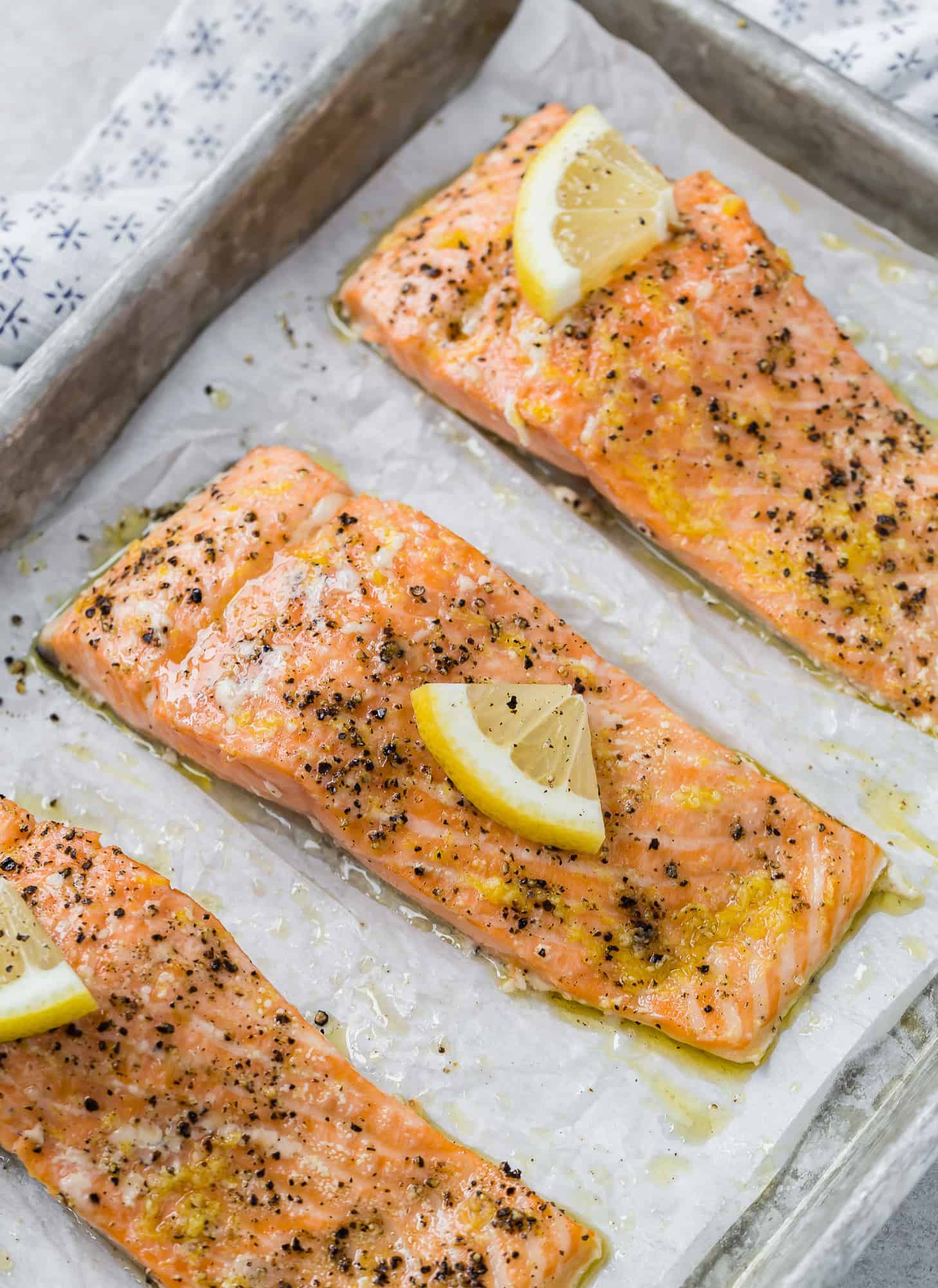 Three slow roasted salmon filets topped with lemon slices and black pepper on a parchment lined baking sheet.