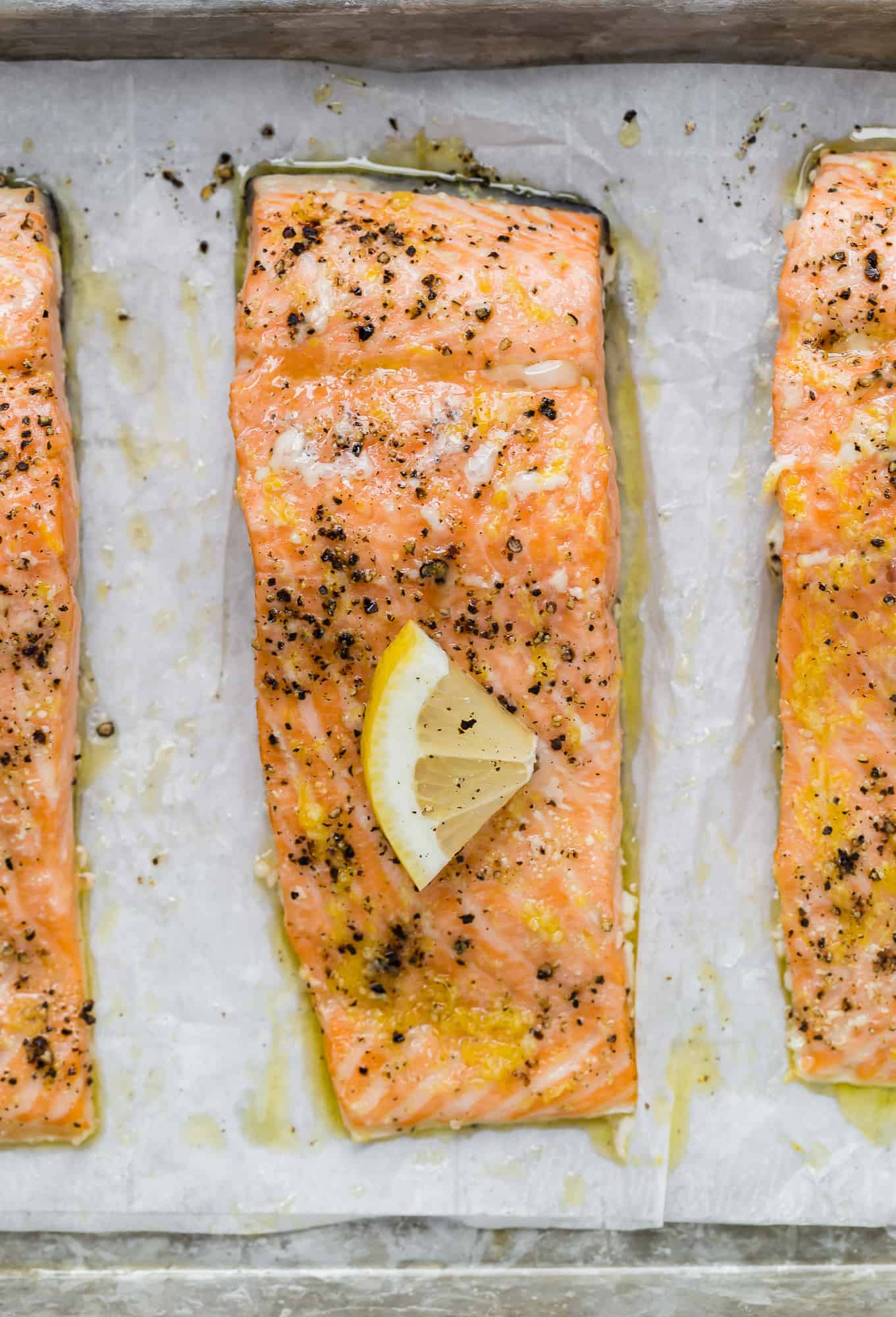 Close up overhead view of salmon filets on a baking sheet topped with lemon slices and black pepper.