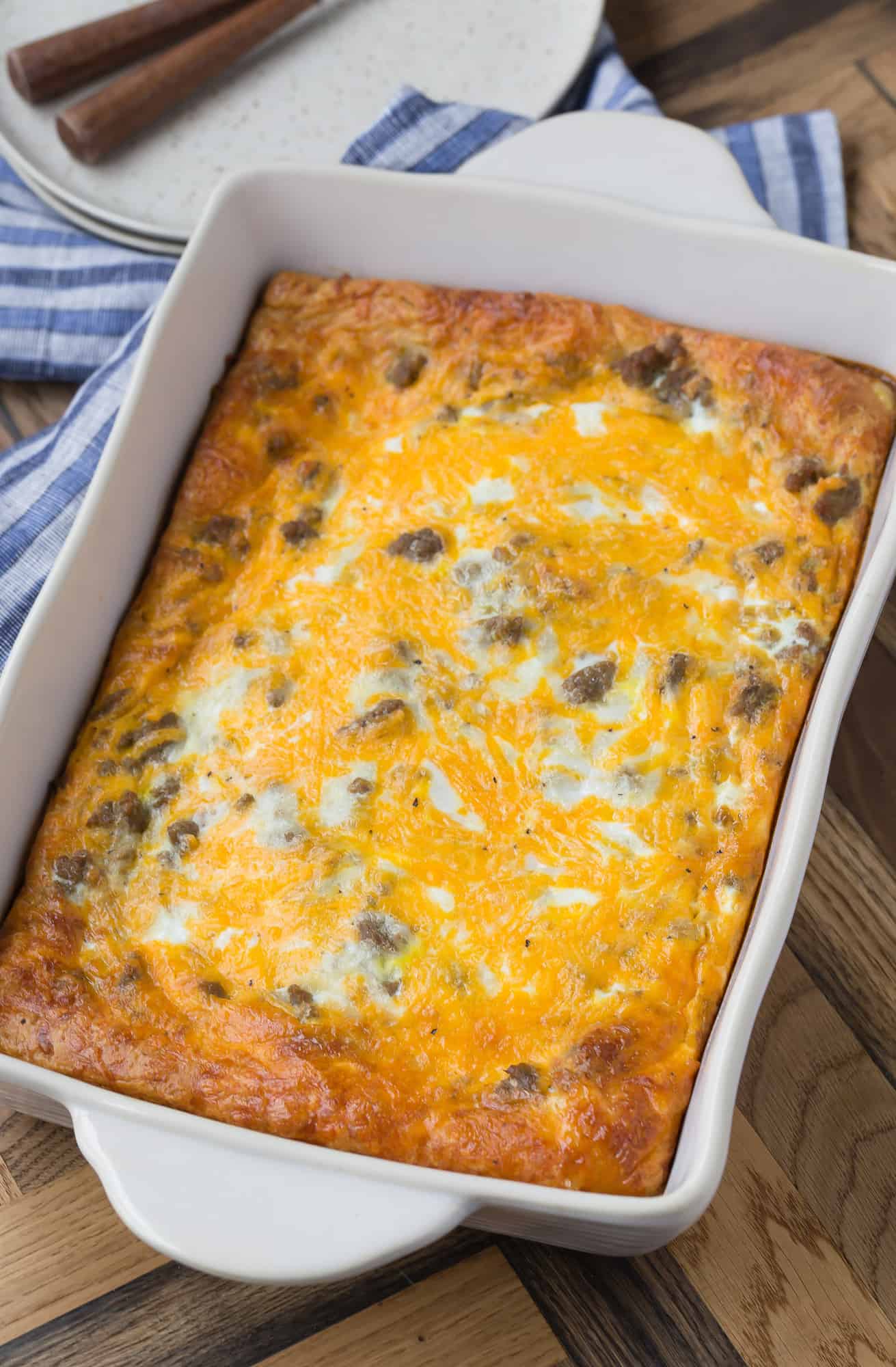 A white 9x13 baking dish on a wooden background. Dish is full of a cheese and sausage breakfast casserole. Forks and plates are partially visible in the background.