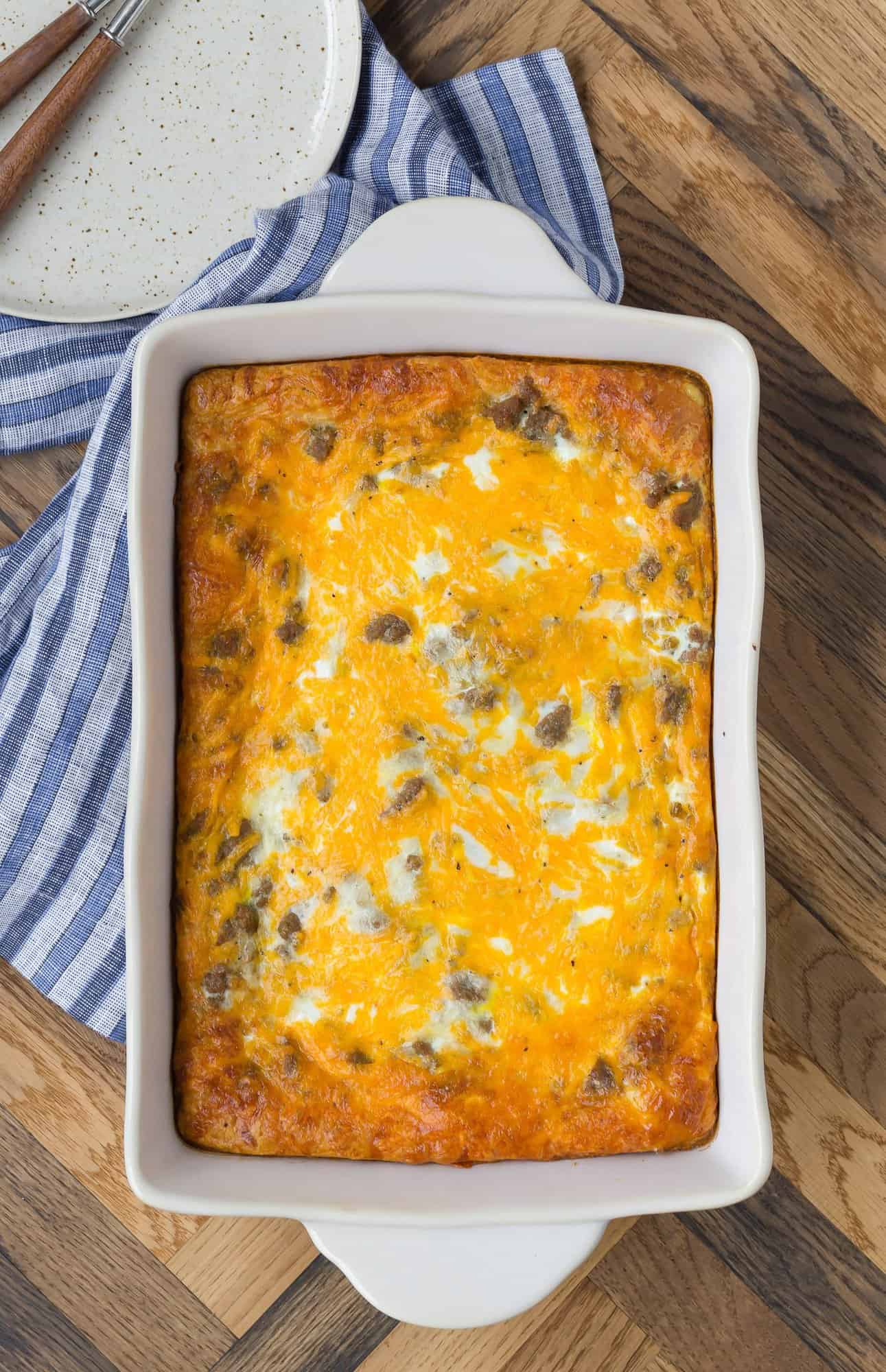 Overhead view of a pan of cheesy breakfast casserole on a wooden backdrop. Plates, forks, and a striped linen are also pictured.