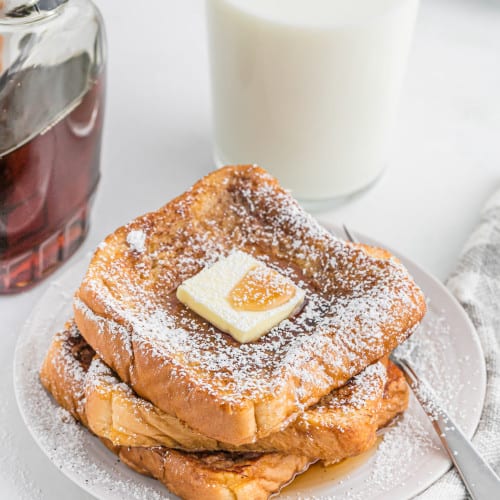 French toast stacked on a plate with powdered sugar, butter, and maple syrup.
