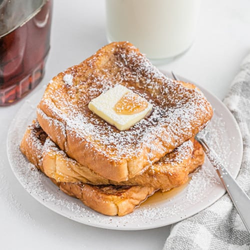 French toast stacked three high on a white plate, topped with a pat of butter and powdered sugar.