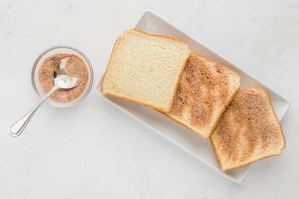 Bread slices on a plate dusted with cinnamon and sugar, next to a small bowl of cinnamon sugar with a spoon.