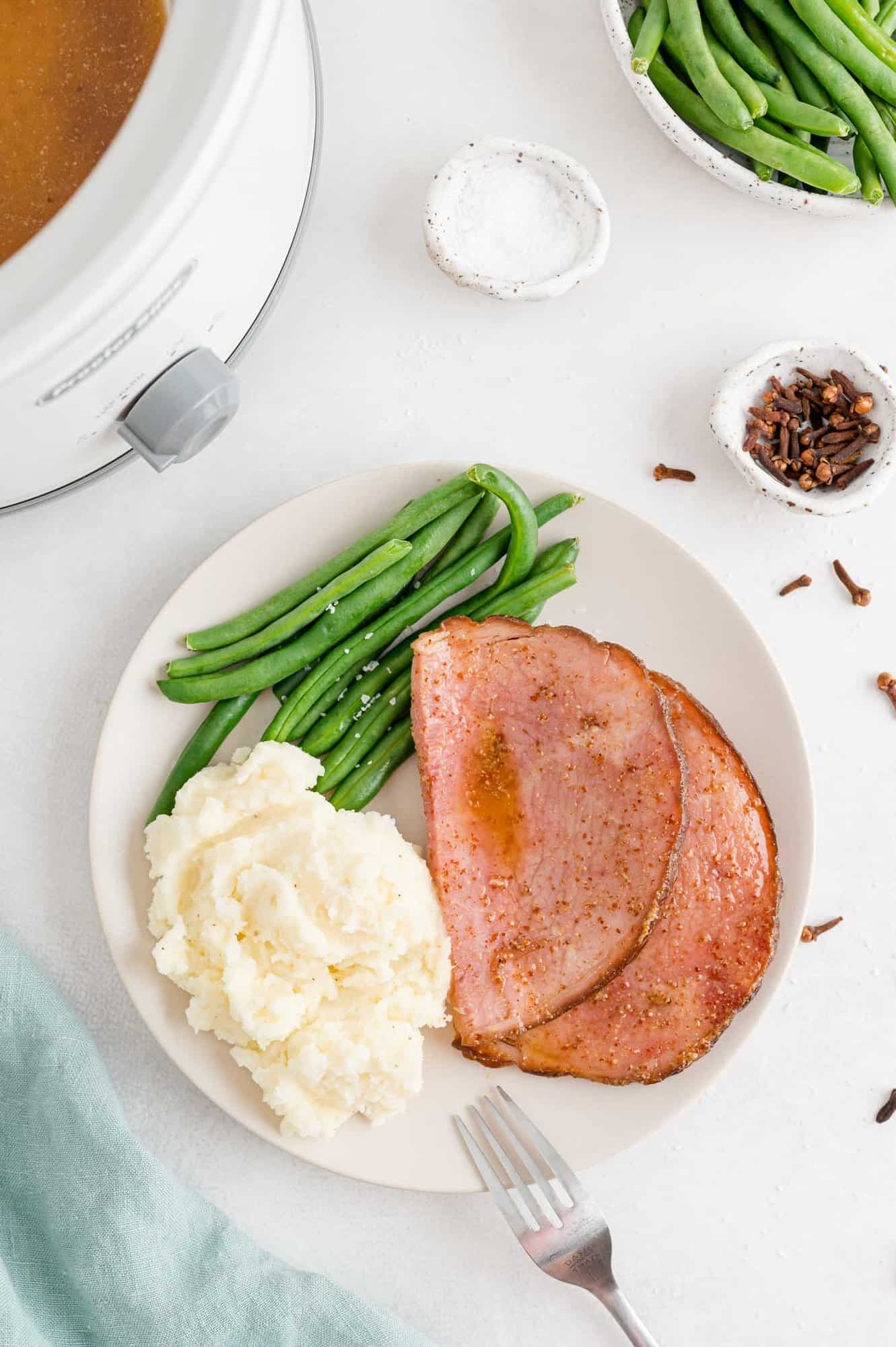 Crockpot ham on a plate with mashed potatoes and green beans.