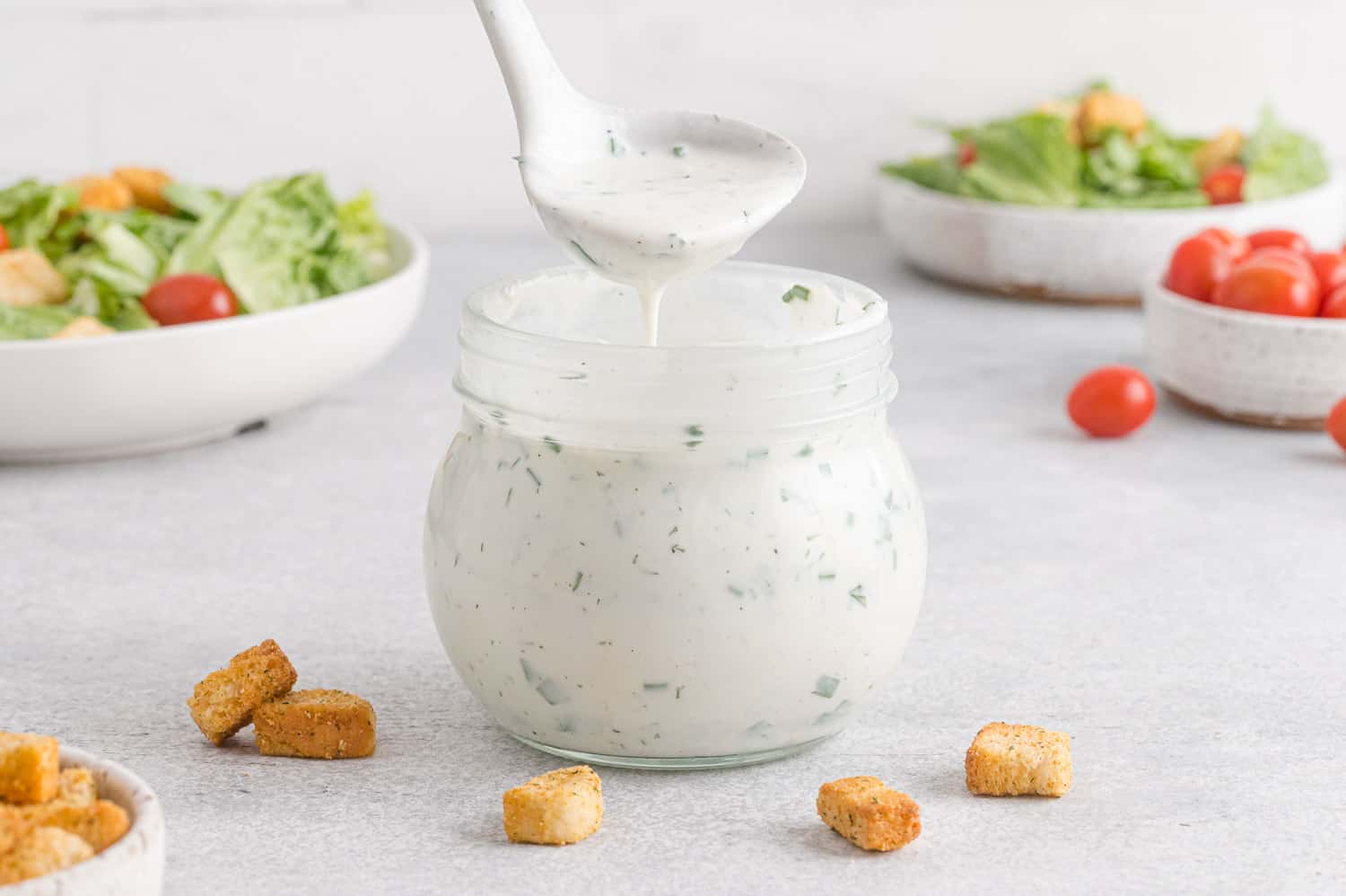 Ranch dressing shown on a ladle and in a jar, salads in background.