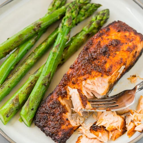 Cooked salmon fillet on a grey-toned plate with cooked asparagus spears. The salmon is being flaked with a fork.