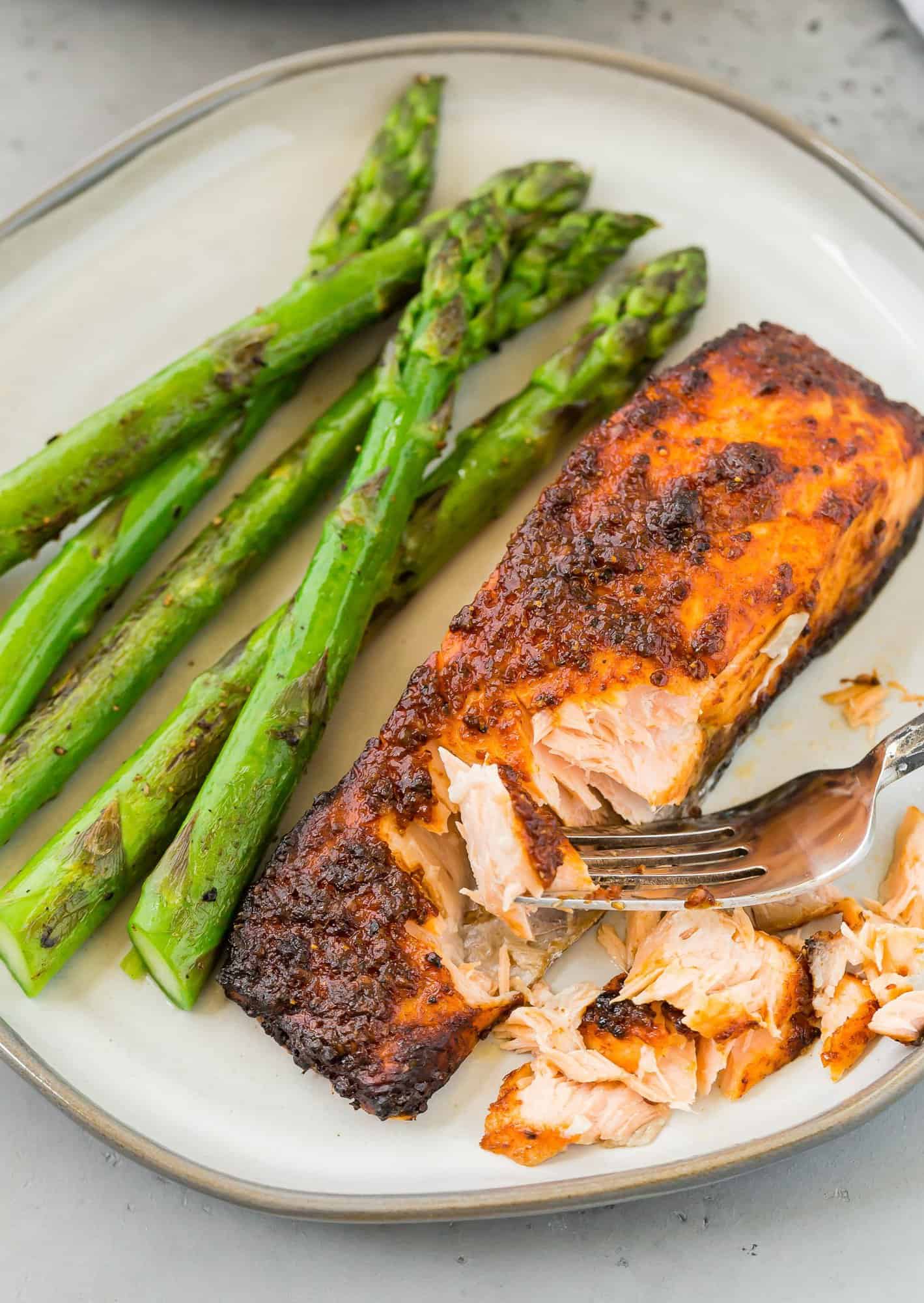 Cooked salmon fillet on a grey-toned plate with cooked asparagus spears. The salmon is being flaked with a fork.