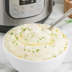 A bowl of mashed potatoes garnished with chopped chives, with a spoon in it, placed in front of an Instant Pot on a marble countertop.
