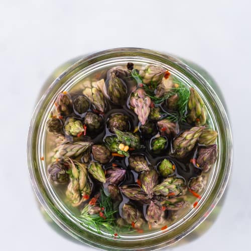 Pickled asparagus in a jar, photographed from above, showing the flowery ends of the asparagus spears.