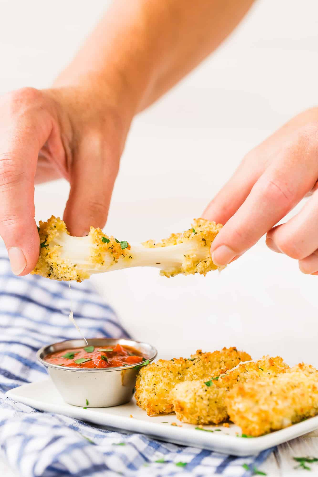 Air fried mozzarella stick, being pulled apart with a large sting of mozzarella cheese connecting the two halves. Additional mozzarella sticks are pictured along with a small cup of marinara sauce.