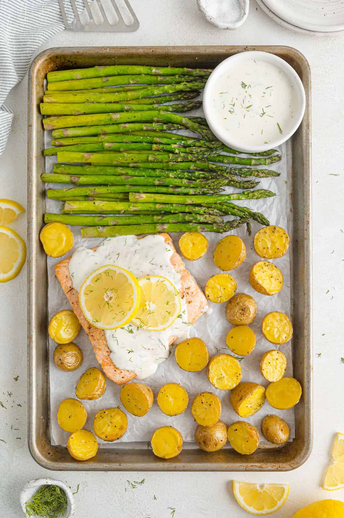 Overhead view of a salmon sheet pan dinner, with roasted asparagus and potatoes next to a salmon filet topped with lemon dill sauce on a baking pan.