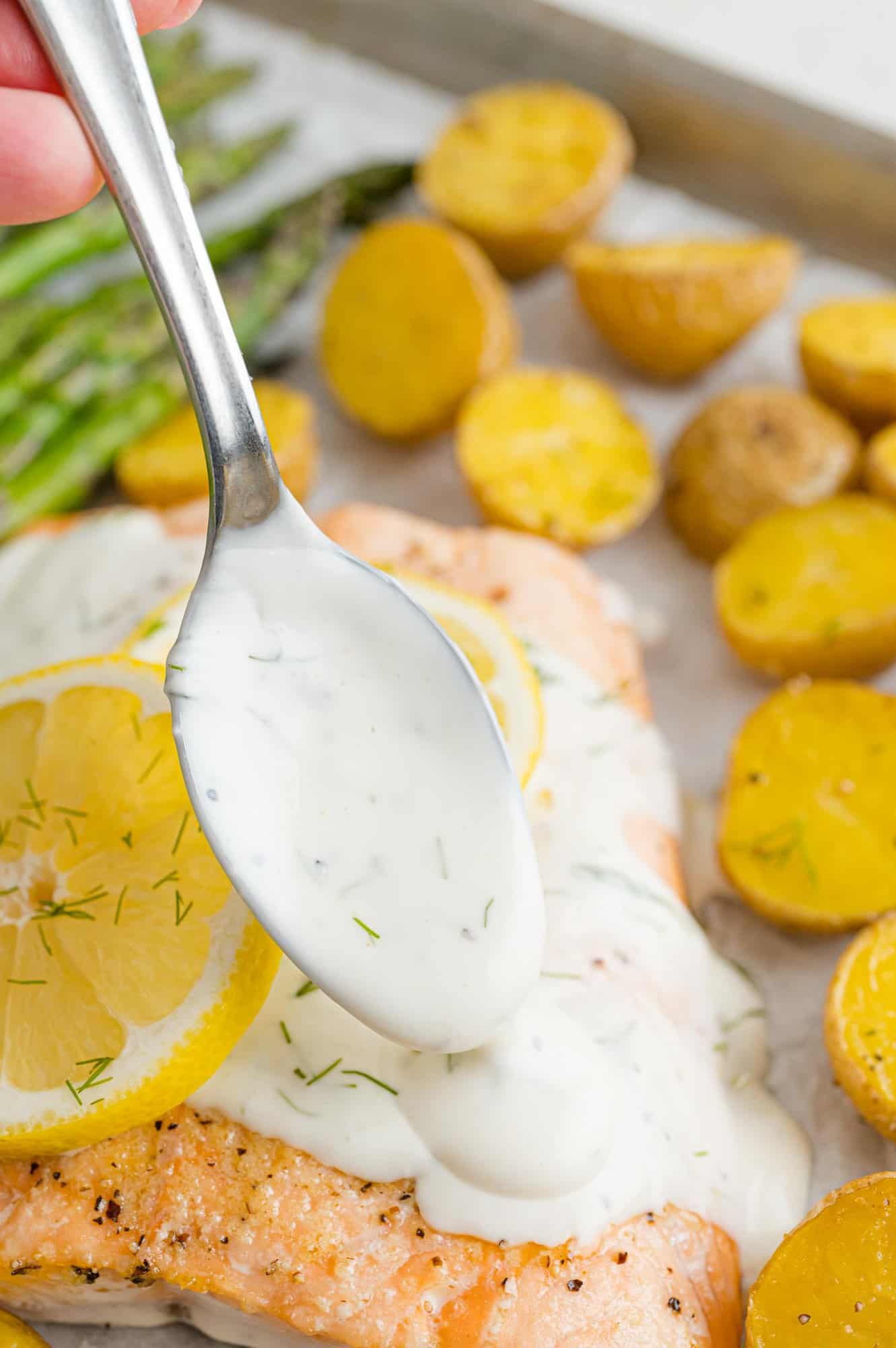 A spoon drizzles lemon dill sauce over a salmon filet on a sheet pan next to roasted potatoes and asparagus.