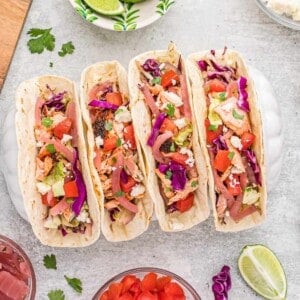 Overhead view of a row of salmon tacos with garnishes in a taco holder, surrounded by taco toppings.