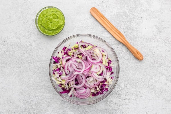 A small bowl of avocado dressing next to a bowl of shredded red cabbage and onions.