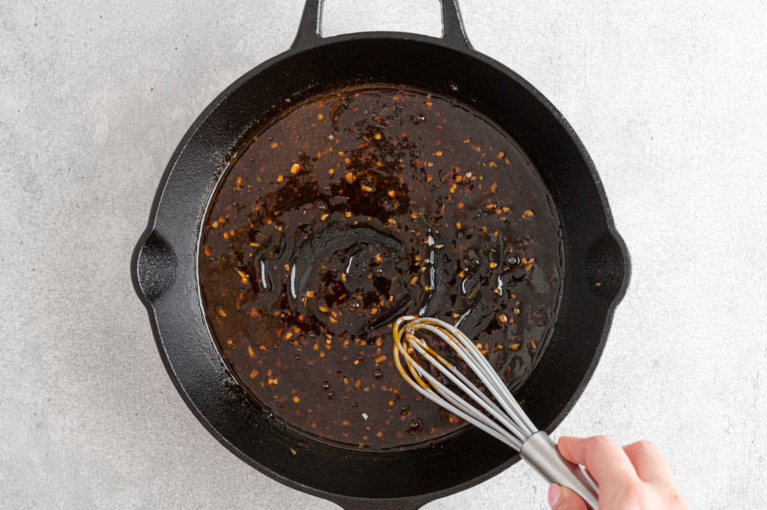 A hand using a whisk to stir glaze as it thickens in pan.