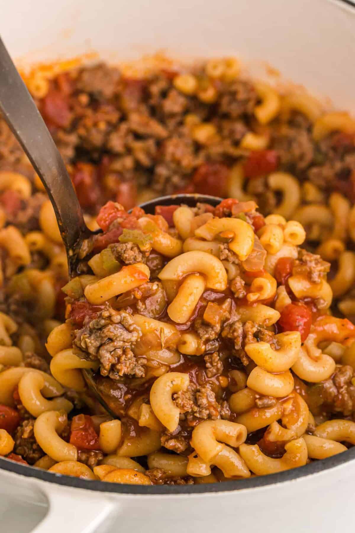 A ladle scoops up American goulash with macaroni, ground beef, tomatoes, and sauce from a large pot.