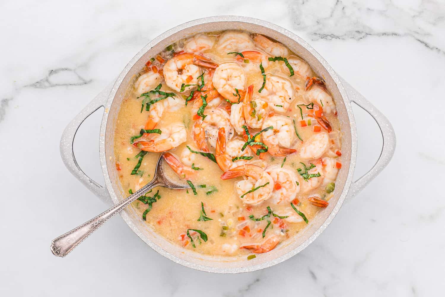 A pot of shrimp in a creamy sauce with chopped herbs and diced vegetables, served with a metal spoon on a marble surface.