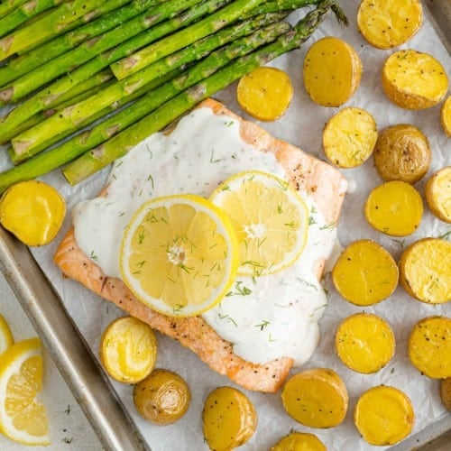 Overhead view of salmon, potatoes, and asparagus on a sheet pan topped with lemon dill sauce.