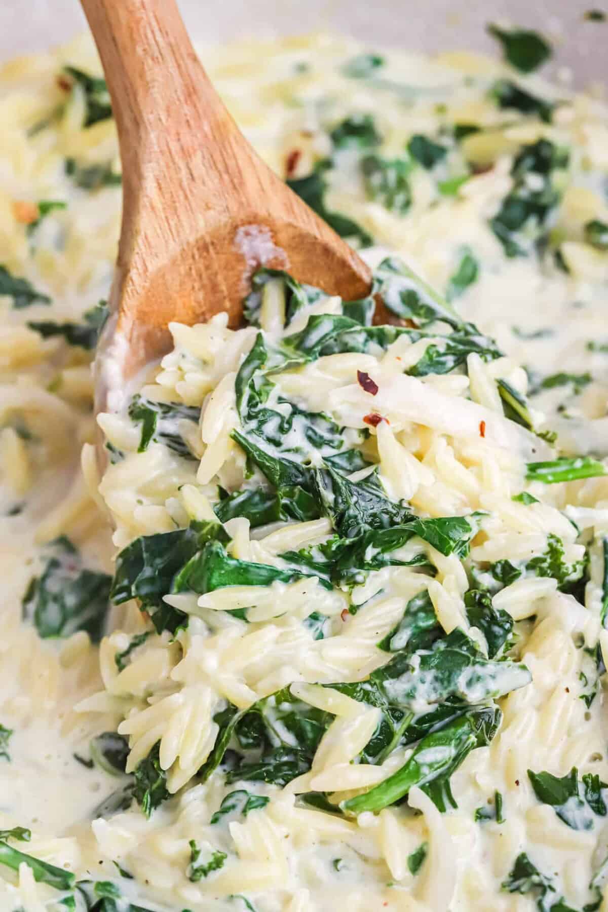 A wooden spoon stirs creamy orzo pasta with wilted spinach and a white sauce.