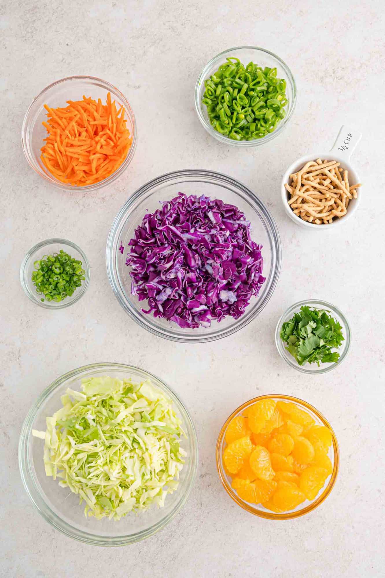 Assorted ingredients in glass bowls including shredded red and green cabbage, mandarin orange segments, shredded carrot, green onions, snap peas, cilantro, and crunchy noodles.